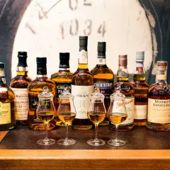 Rząd butelek szkockiej whisky, od Dalwhinnie po Monkey Shoulder, z kieliszkami pełnymi bursztynowego płynu. Czy to początek dyskusji: co jest lepsze koniak czy whisky?
