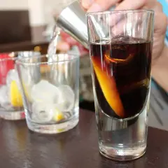 Przygotowanie drinków. W szklance ciemny płyn z plasterkiem pomarańczy i lodem. Obok szklanki z lodem i cytryną.