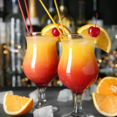 Dwa kolorowe, domowe drinki z wódką, udekorowane pomarańczą i wiśnią, z kostkami lodu i owocami w tle.