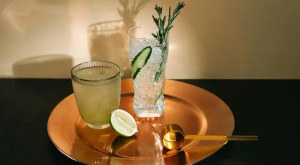 Orzeźwiający drink z limonką, lodem i rozmarynem. Obok drugi napój i złota miarka.