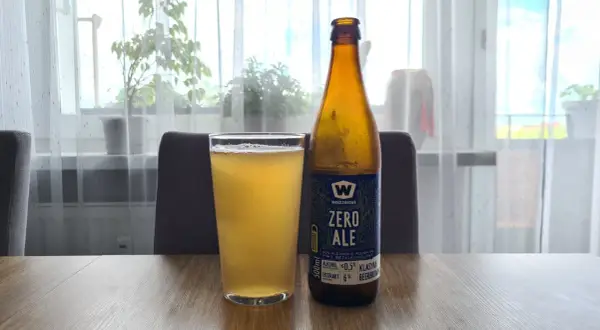 Szklanka i butelka piwa bezalkoholowego "Zero Ale" na drewnianym stole. Etykieta informuje, co zawiera piwo bezalkoholowe: ekstrakt 6%, alkohol ≤0.5%.