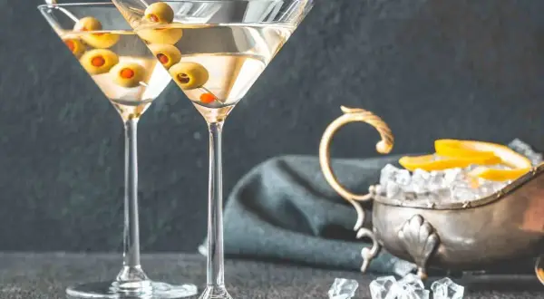 Dwa kieliszki z martini, ozdobione oliwkami, czekają na degustację. Obok lód i cytryna, idealne dodatki, by dowiedzieć się, jak pić martini.