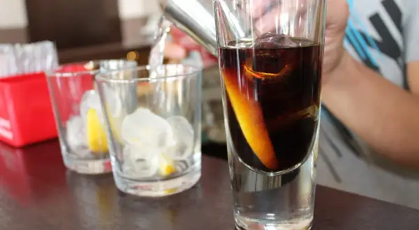 Przygotowanie drinków. W szklance ciemny płyn z plasterkiem pomarańczy i lodem. Obok szklanki z lodem i cytryną.