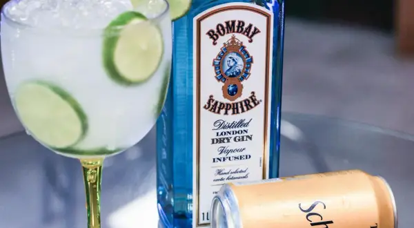 Orzeźwiający drink z tonikiem i limonką, obok butelka ginu Bombay Sapphire i puszka Schweppes. Idealny na letnie wieczory.