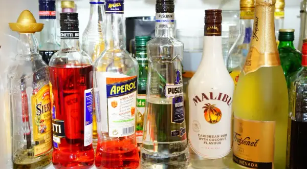 Na półce stoją butelki z różnymi alkoholami. Wśród nich wódka Puschkin, która ma 40% alkoholu, Aperol, Malibu i Freixenet.