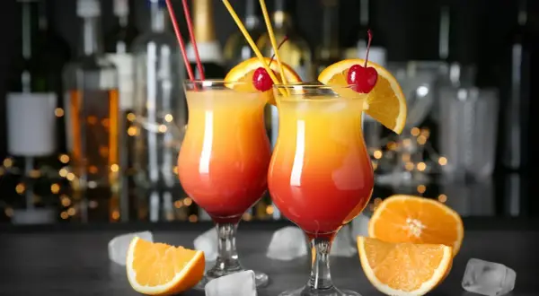 Dwa kolorowe, domowe drinki z wódką, udekorowane pomarańczą i wiśnią, z kostkami lodu i owocami w tle.