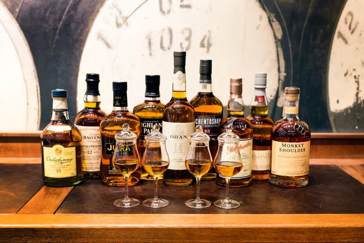 Rząd butelek szkockiej whisky, od Dalwhinnie po Monkey Shoulder, z kieliszkami pełnymi bursztynowego płynu. Czy to początek dyskusji: co jest lepsze koniak czy whisky?