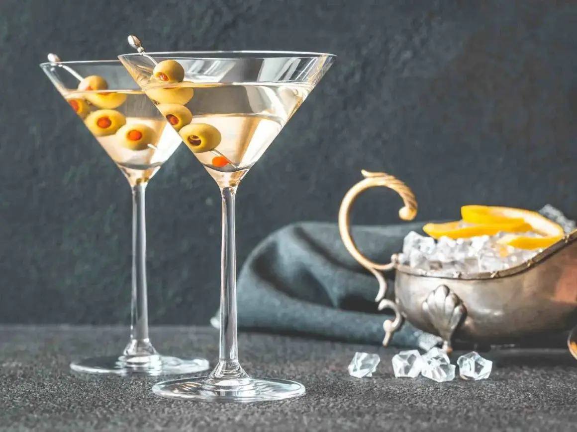 Dwa kieliszki z martini, ozdobione oliwkami, czekają na degustację. Obok lód i cytryna, idealne dodatki, by dowiedzieć się, jak pić martini.
