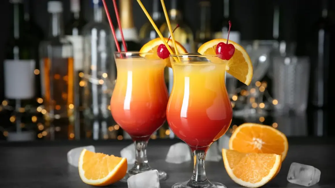 Dwa kolorowe, domowe drinki z wódką, udekorowane pomarańczą i wiśnią, z kostkami lodu i owocami w tle.