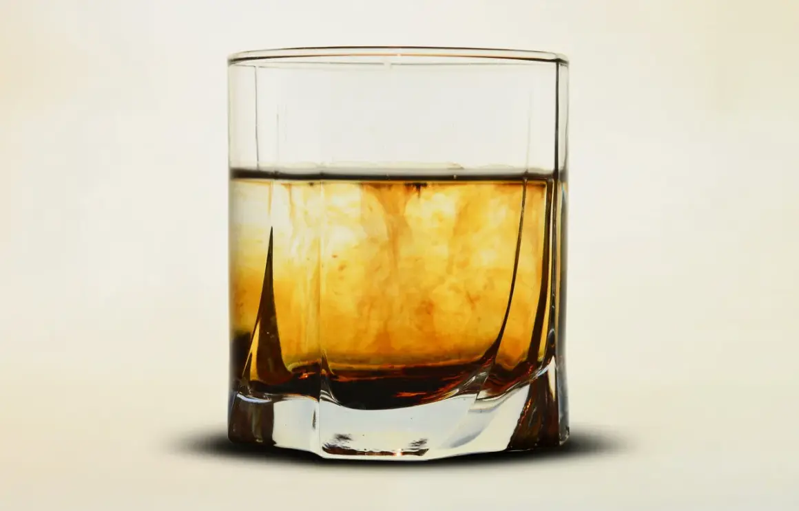 Szklanka z bursztynowym płynem, w którym rozpuszcza się barwnik. Wizualizacja procesu, jak zrobić whisky w domu.