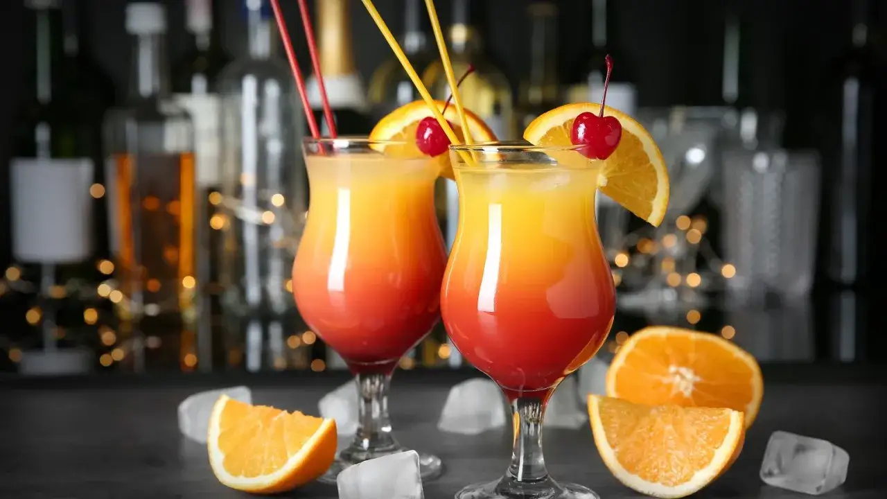 Dwa kolorowe, domowe drinki z wódką, udekorowane pomarańczą i wiśnią, z kostkami lodu i owocami w tle.