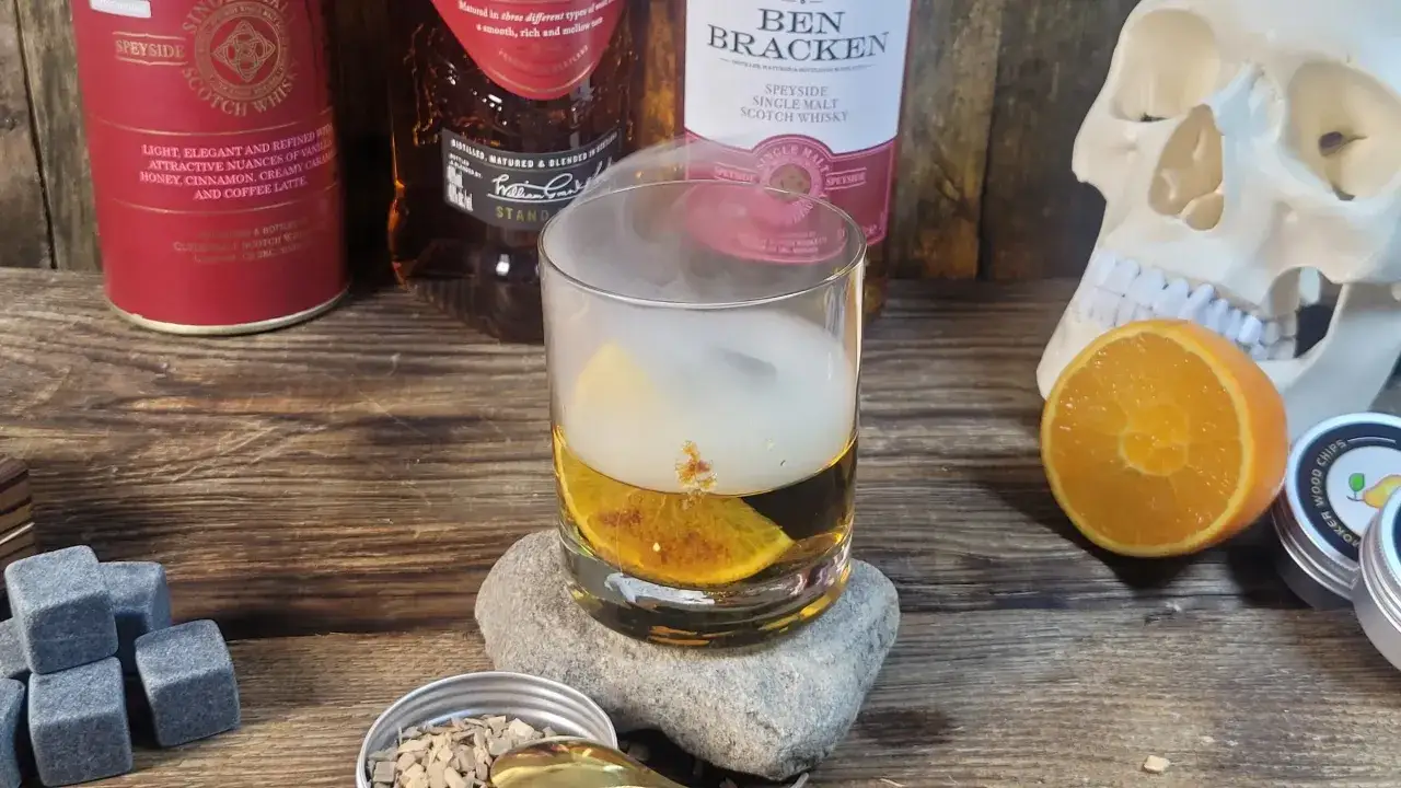 Dym unoszący się nad szklanką whisky z pomarańczą. Czy whisky podnosi cukier? Obok butelki, czaszka i kamienne kostki.