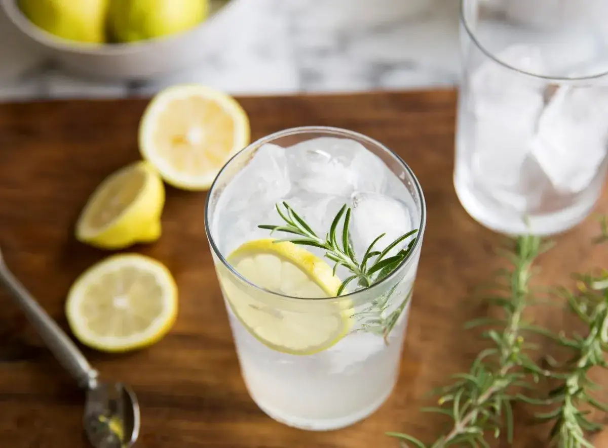 Orzeźwiający drink z tonikiem, lodem, plasterkiem cytryny i gałązką rozmarynu. Obok leżą cytryny i puste szklanki.