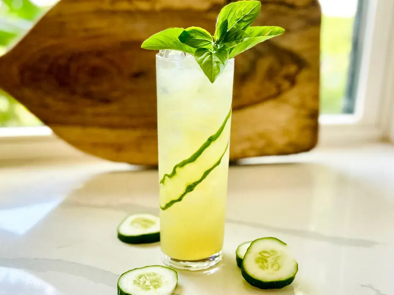 Orzeźwiający drink z limonką, ozdobiony plastrami ogórka i listkami bazylii, na tle drewnianej deski.